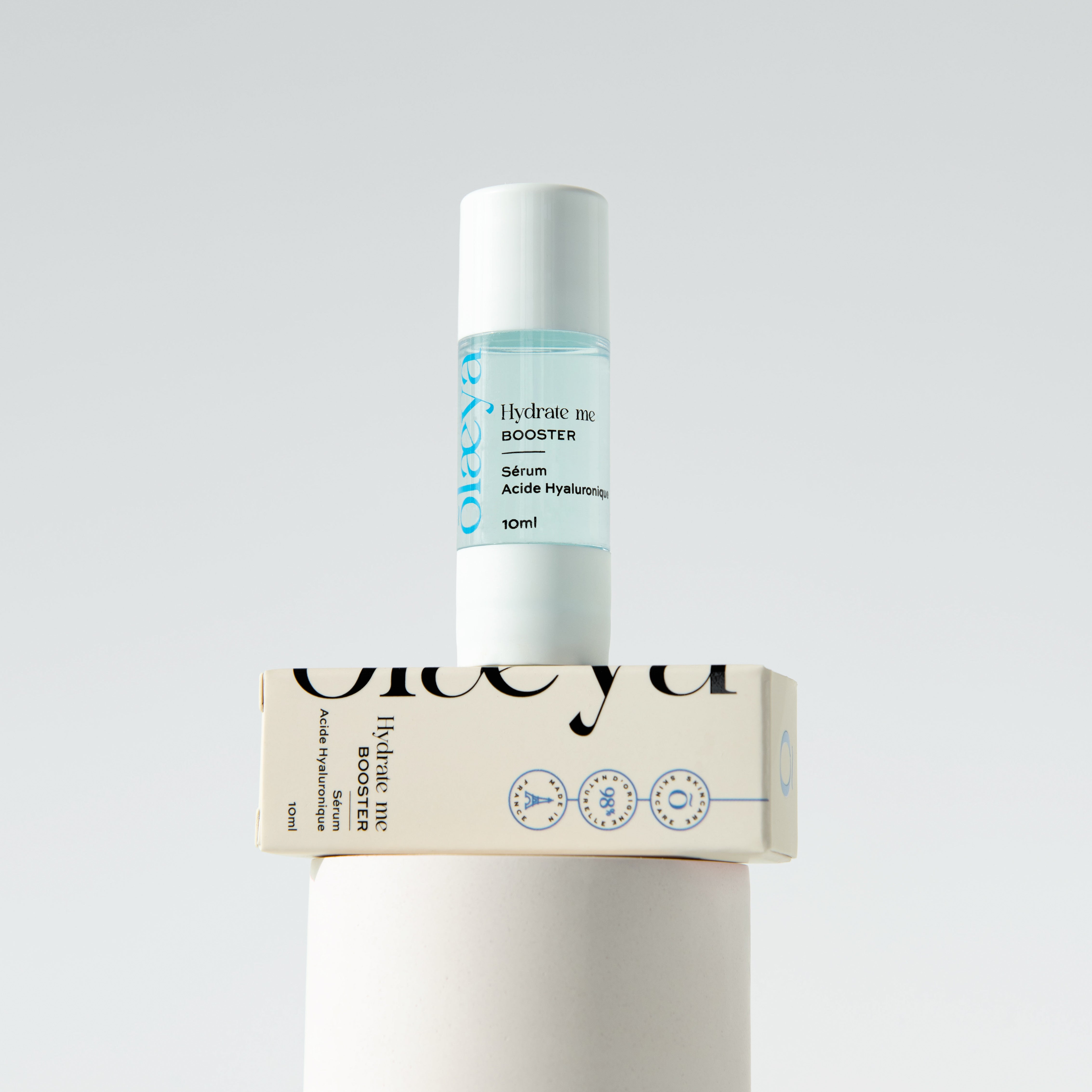 Flacon 10 ml du sérum à l'acide hyaluronique Booster Hydrate Me Olaeya posé sur sa boîte, fond gris